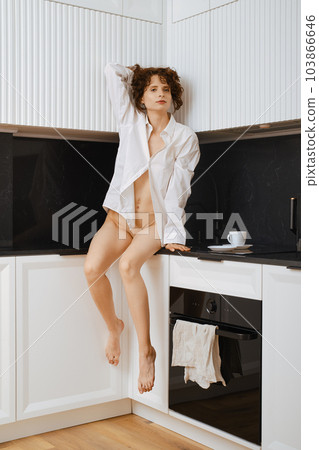 Carefree woman with tousled hair in shirt over her naked body sits in kitchen 103866646