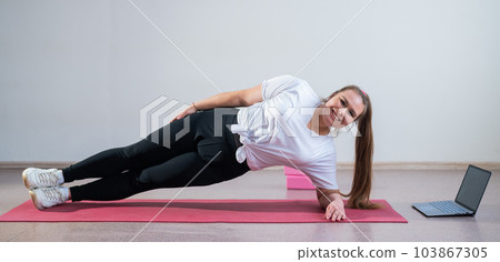 Young fat caucasian woman doing a side plank on a white background. A cute plus size girl in sportswear is doing fitness exercises and watching an online tutorial on a laptop 103867305