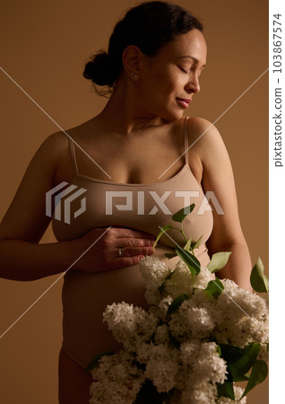 Pregnancy. Maternity. Expecting baby. Multiethnic attractive delightful young pregnant woman in beige lingerie, holding hand on her belly, posing with blooming white lilacs, isolated studio background Pregnancy. Maternity. Expecting baby. Multiethnic attractive delightful young pregnant woman in beige lingerie, holding hand on her belly, posing with blooming white lilacs, isolated studio background 103867574