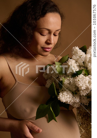 Close-up studio portrait of beautiful multiethnic gravid woman, pregnant expectant mother in beige lingerie, holding bouquet of blooming lilacs, isolated background. Pregnancy fashion. Body positivity Close-up studio portrait of beautiful multiethnic gravid woman, pregnant expectant mother in beige lingerie, holding bouquet of blooming lilacs, isolated background. Pregnancy fashion. Body positivity 103867576