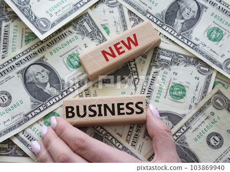 New business symbol. Concept words New business on wooden blocks. Businessman hand. Beautiful dollar background. Business and New business concept. Copy space. New business symbol. Concept words New business on wooden blocks. Businessman hand. Beautiful dollar background. Business and New business concept. Copy space. 103869940