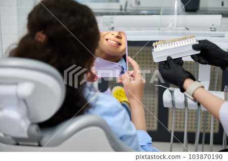 Rear view woman in dental chair, smiles looking at mirror, while a dentist using tooth color chart to compare with patient's teeth, preparing for laser teeth whitening bleaching in dentistry clinic. 103870190