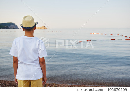 Teenage boy in a straw hat and a white t-shirt looks into the sea. Concept of acation and travel. Teenage boy in a straw hat and a white t-shirt looks into the sea. Concept of acation and travel. 103870309