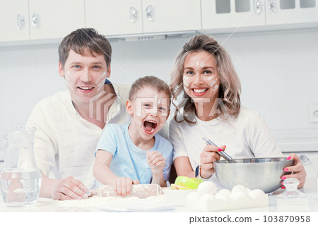 Young family with child is sitting at kitchen table covered in flour and laughing merrily. Portrait Young family with child is sitting at kitchen table covered in flour and laughing merrily. Portrait 103870958