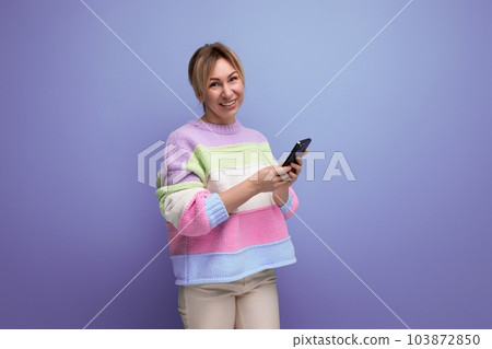 blond smiling young woman in casual outfit holding a smartphone in her hands on a purple background 103872850