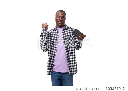 positive smiling young african man with short haircut in plaid shirt rejoices in victory holding 103873042