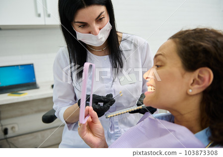 Female patient sitting in dentists chair, smiles looking at mirror while visiting dentist for teeth bleaching procedure. Teeth prosthesis. Teeth bleaching or whitening concept. Dental practice Female patient sitting in dentists chair, smiles looking at mirror while visiting dentist for teeth bleaching procedure. Teeth prosthesis. Teeth bleaching or whitening concept. Dental practice 103873308