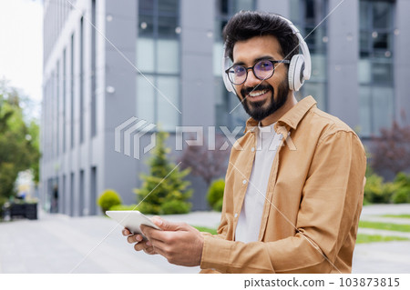 Portrait young Indian man with headphones and tablet watching online video sitting on bench near office building businessman smiling and looking at camera, portrait of satisfied online stream viewer Portrait young Indian man with headphones and tablet watching online video sitting on bench near office building businessman smiling and looking at camera, portrait of satisfied online stream viewer 103873815