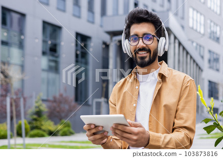 Portrait young Indian man with headphones and tablet watching online video sitting on bench near office building businessman smiling and looking at camera, portrait of satisfied online stream viewer Portrait young Indian man with headphones and tablet watching online video sitting on bench near office building businessman smiling and looking at camera, portrait of satisfied online stream viewer 103873816