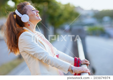smiling trendy woman in pink dress and white jacket in city 103874974