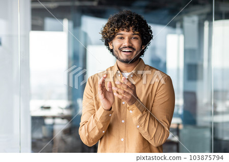 Businessman standing by the window applauding to the camera to colleagues, video call online meeting, young Latin American smiling and looking at the web. Businessman standing by the window applauding to the camera to colleagues, video call online meeting, young Latin American smiling and looking at the web. 103875794