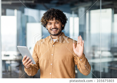 Portrait of young programmer with tablet computer inside office, hispanic man with curly hair smiling and looking at camera, man at workplace testing online applications standing near window. Portrait of young programmer with tablet computer inside office, hispanic man with curly hair smiling and looking at camera, man at workplace testing online applications standing near window. 103875803