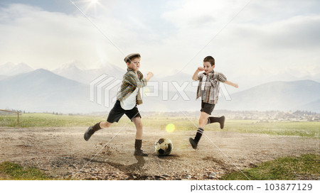 Two little boys, children in retro style clothes playing football outdoors on a daytime. Countryside and mountains background Two little boys, children in retro style clothes playing football outdoors on a daytime. Countryside and mountains background 103877129