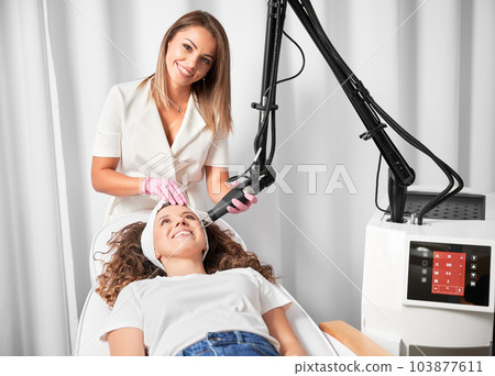 Female doctor cosmetologist using erbium ablative laser machine while performing resurfacing procedure. Smiling woman lying on daybed and receiving facial skincare treatment in beauty salon. 103877611