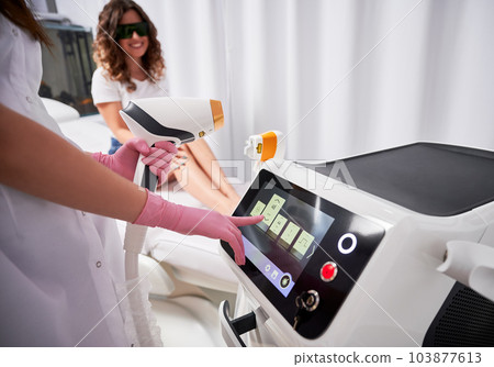 Close-up view hands of beautician in pink gloves, who holding laser sensor with one hand and adjusting laser device on scoreboard with the other. Satisfied client in the blurred background. 103877613