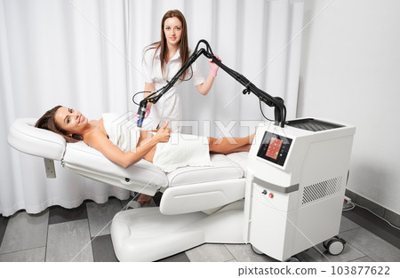 Smiling client lying on white couch and showing class during procedure of abdomen skin laser resurfacing in beauty salon. Beautiful young woman satisfied with skin rejuvenation procedure. 103877622