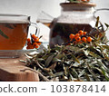 The concept of medicinal herbal tea. A pile of dry sea buckthorn leaves prepared for medicinal tea against the background of a teapot and a cup of tea. 103878414