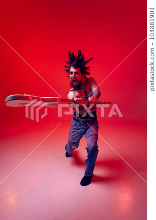 Crazy punk. Emotional young man, rock musician posing aggressively with electric guitar against red studio background in neon light 103881901