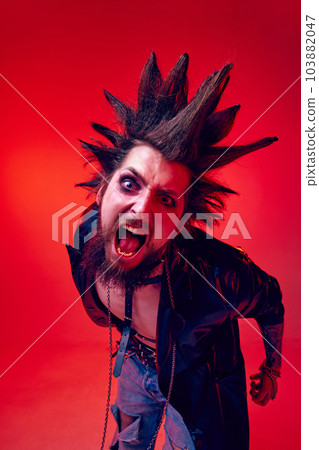 Crazy face. Portrait of expressive young man, punk posing in makeup and leather jacket against red studio background in neon light Crazy face. Portrait of expressive young man, punk posing in makeup and leather jacket against red studio background in neon light 103882047