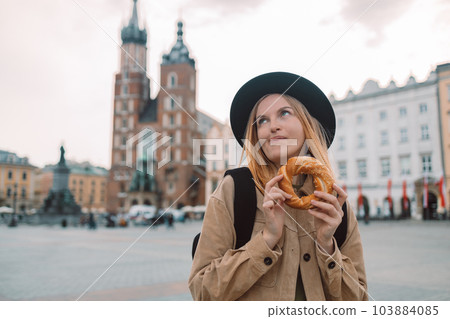 Attractive young female tourist with stylish clothes and a boho hat eating bagel obwarzanek traditional polish cuisine snack waling on Market square in Krakow. Traveling Europe Attractive young female tourist with stylish clothes and a boho hat eating bagel obwarzanek traditional polish cuisine snack waling on Market square in Krakow. Traveling Europe 103884085