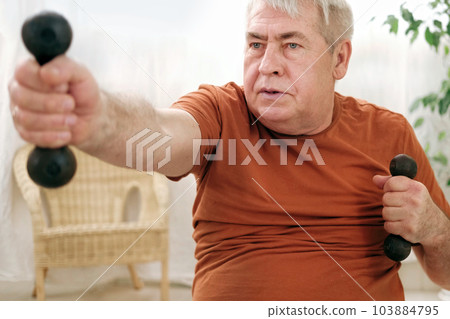 Old elderly man in sportswear training in living room doing weight lifting dumbbell exercising at home. Leisure healthy lifestyle. Older male is working out. Fitness for seniors. Active retired people Old elderly man in sportswear training in living room doing weight lifting dumbbell exercising at home. Leisure healthy lifestyle. Older male is working out. Fitness for seniors. Active retired people 103884795