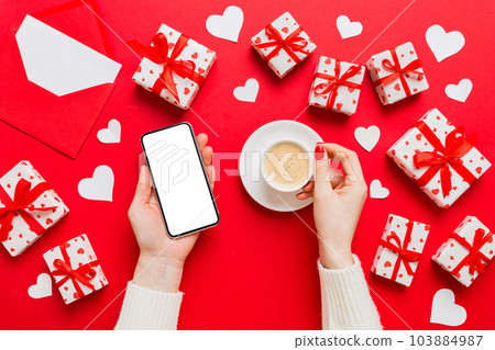 Female hands holding smart phone with coffee for Valentine day, gift box and envelope, hearts Female hands holding smart phone with coffee for Valentine day, gift box and envelope, hearts 103884987