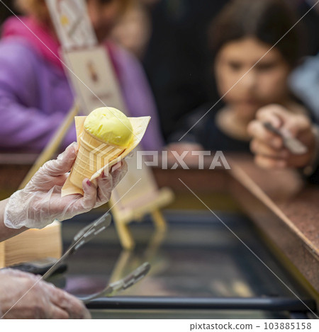 Hand Of Vendor Holding fruit Ice Cream Cone, background of counter with sweet dessert. Real scene in store Hand Of Vendor Holding fruit Ice Cream Cone, background of counter with sweet dessert. Real scene in store 103885158