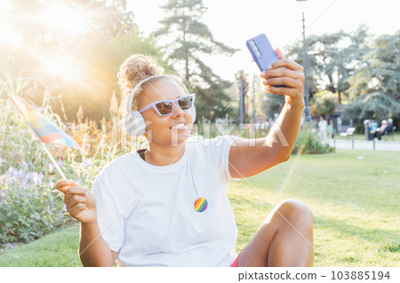Young Latin happy woman wrapped in a rainbow flag, symbol of LGBT community, makes selfie on a mobile phone on a pride celebration in a city park. Human Rights, Equality, LGBT, Freedom and Happiness Young Latin happy woman wrapped in a rainbow flag, symbol of LGBT community, makes selfie on a mobile phone on a pride celebration in a city park. Human Rights, Equality, LGBT, Freedom and Happiness 103885194