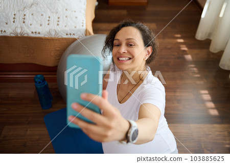 Beautiful pregnant woman sitting on blue exercise mat at home, smiling broadly taking selfie on smart mobile phone, sharing her happy moments of her pregnancy and maternity lifestyle in social media 103885625