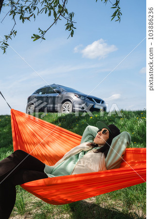 A woman in a cap is lying in a hammock and enjoying nature with a car behind her 103886236