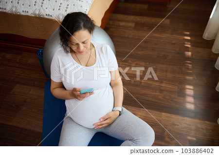 Overhead view pregnant woman mother touching her belly sitting on exercise mat, checking pregnancy mobile app on her smartphone, controlling heart rate and pulse after prenatal fitness workout at home Overhead view pregnant woman mother touching her belly sitting on exercise mat, checking pregnancy mobile app on her smartphone, controlling heart rate and pulse after prenatal fitness workout at home 103886402