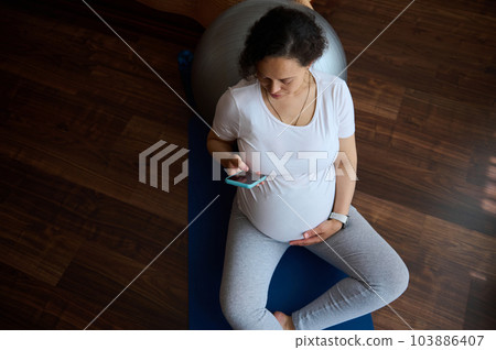 Overhead view of a pregnant woman using smartphone on exercise mat after prenatal fitness at home. Gravid expectant female browsing mobile application, resting after meditation practice. Pregnancy 103886407