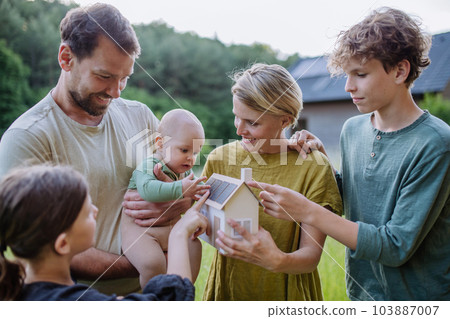 Happy family with three children holding model of house ith solar photovoltaics. Alternative energy, saving resources and sustainable lifestyle concept. 103887007
