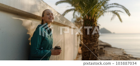 Young fitness woman enjoying coffee after workout, resting at the walkway along the shore. Young fitness woman enjoying coffee after workout, resting at the walkway along the shore. 103887034