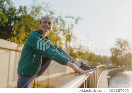 Portrait of young female runner stretching legs before her early morning run along the embankment of river. 103887035