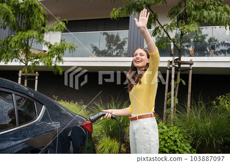 Close up of beautiful woman waving, greeting someone while charging her electric car on the street. Close up of beautiful woman waving, greeting someone while charging her electric car on the street. 103887097
