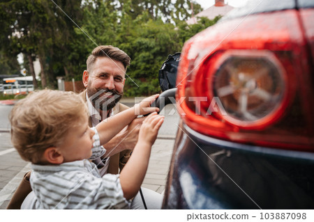 Close up of mature man and his little son charging their electric car on the street. Close up of mature man and his little son charging their electric car on the street. 103887098
