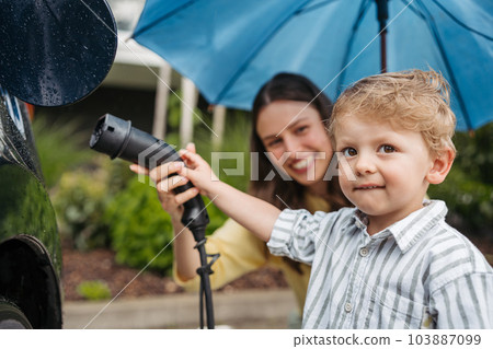 Close up of smiling mother and her little son charging their electric car on a rainy day, standing under umbrella. 103887099