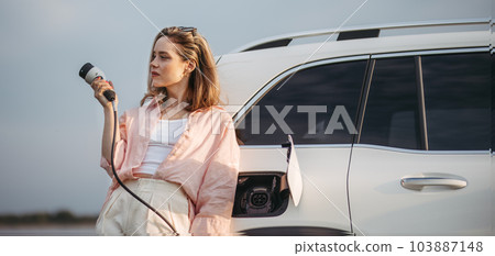 Young woman holding power supply cable from her electric car, prepared for charging it sustainable and economic transportation concept. 103887148