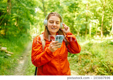 Happy beautiful woman listening music on headphones in the nature in the forest, carrying a backpack in the forest on sunset light in the summer season. Happy beautiful woman listening music on headphones in the nature in the forest, carrying a backpack in the forest on sunset light in the summer season. 103887696