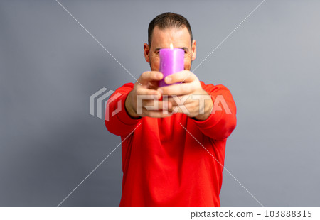 Hispanic man in his 40s wearing a red jumper with a lit purple candle in front of his face, isolated on a gray studio background. 103888315