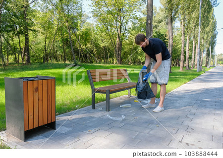 A male volunteer collects plastic waste in a black bag in a public park. 103888444