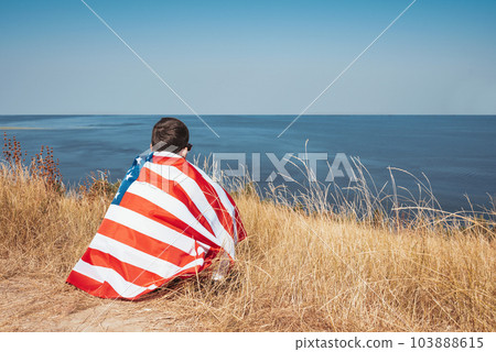 Teenager sits with the American flag on the bay outdoors. Back view Teenager sits with the American flag on the bay outdoors. Back view 103888615