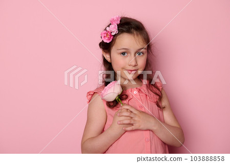 Authentic Caucasian child, beautiful elegant little girl in pink dress, holding a rose flower, smiling cutely looking at camera, isolated pink background. Copy space. Kids beauty and fashion portrait 103888858