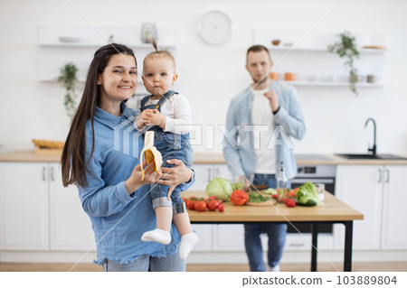 Baby eating banana on mommy's hands while dad making dinner Baby eating banana on mommy's hands while dad making dinner 103889804