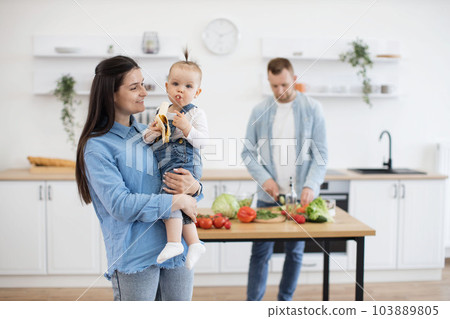 Baby eating banana on mommy's hands while dad making dinner Baby eating banana on mommy's hands while dad making dinner 103889805
