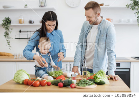 Infant girl mixing salad with metal spoons and mother's help Infant girl mixing salad with metal spoons and mother's help 103889854