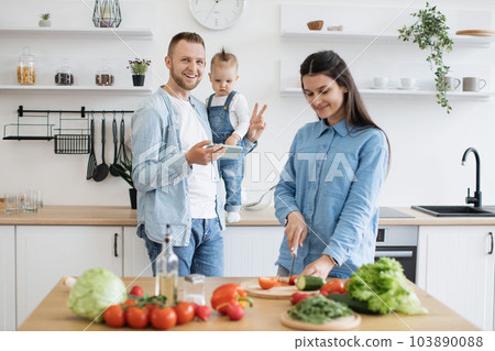 Dad with mobile holding child while mom making homemade dish Dad with mobile holding child while mom making homemade dish 103890088