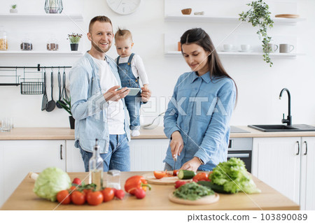 Dad with mobile holding child while mom making homemade dish 103890089