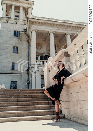 Stylish woman in the city. Fashion photo of a beautiful model in an elegant black dress posing against the backdrop of a building on a city street Stylish woman in the city. Fashion photo of a beautiful model in an elegant black dress posing against the backdrop of a building on a city street 103890463